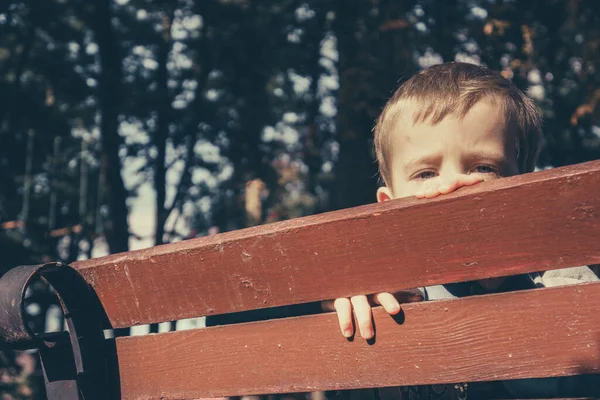 Sad boy hides his face behind a wooden bench in the park