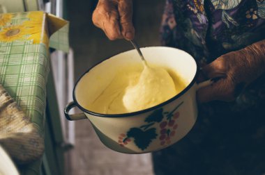 Old grandmother kneads dough for  small pancakes in saucepan in country kitchen, close up