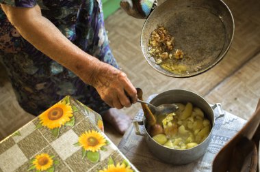 Old woman cooking ground potatoes, putting  roasted onion from pan into aluminum pot