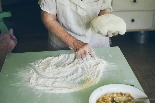 Old woman kneads dough for patties, flour and tureen of stuffing on kitchen table