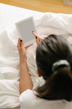 Relaxed young brunette woman reading e-book on bed
