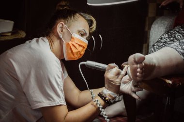 Masked pedicurist grinds dry skin of client's foot