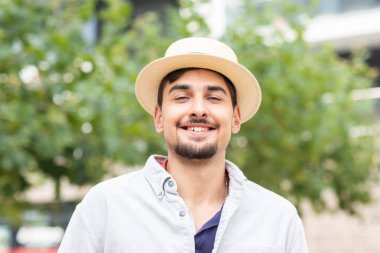 young man with summer hat standing in front of trees in a city
