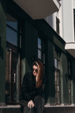 Genç bir kız güzel yeşil bir binanın yanında oturuyor.