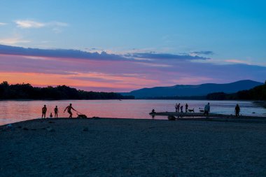 Gün batımında nehir kıyısında insan ve köpeklerin silueti.