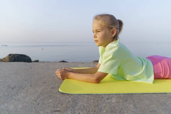 A girl on the beach doing yoga is lying on a yoga mat.