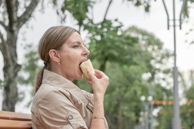 Orta yaşlı bir kadın parkta bankta oturur ve dondurma yer..