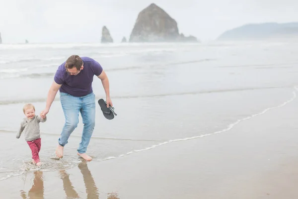 Caucasian father and toddler son run in water in Cannon Beach.