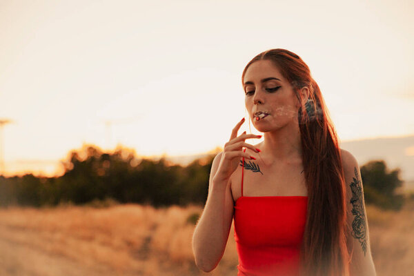 Татуированная девушка в красном платье курит марихуану на закате