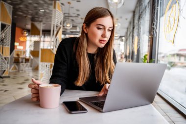 Pencerenin yanındaki kafede dizüstü bilgisayarla çalışan genç bir kız.