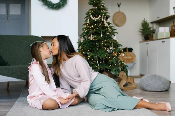 Mother kisses daughter on the nose while sitting by the Christmas tree