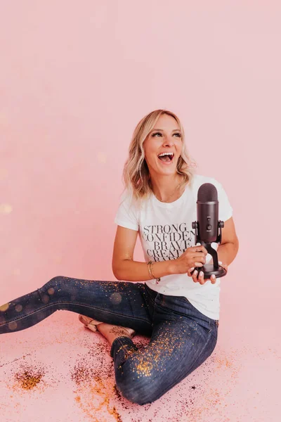 Woman sits on floor covered in glitter holding podcast microphone