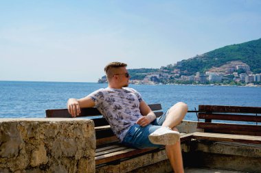 Genç adam sahilde bir bankta oturuyor.      