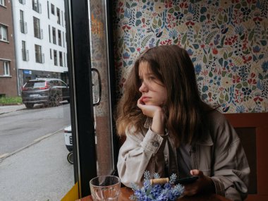 Kafenin penceresinde sakin bir gülümsemeyle oturan genç bir kızın portresi.