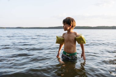 Yüzme pantolonu ve şişme kolları olan 6 yaşındaki bir çocuk gölün suyunda duruyor.