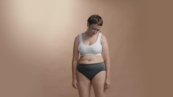 Αναστατωμένη γυναίκα συν μέγεθος βλέποντας τον εαυτό της πιέζοντας την κοιλιά και το λίπος των ποδιών της. Υψηλής ποιότητας μέσο full shot στούντιο βίντεο. — Αρχείο Βίντεο