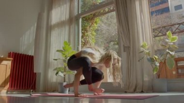 Uzun saçlı, zayıf, spor sütyenli ve taytlı güzel beyaz kadın evde yoga pozu veriyor ve amuda kalkıyor. Güneşli bir gün. 4K video görüntüsü yüklendi