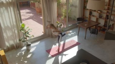 Uzun saçlı, çekici, beyaz, spor sütyenli ve taytlı bir kadın evde yoga pozu veriyor. Güneşli bir gün. En üstteki görünüm 4k video kaydı