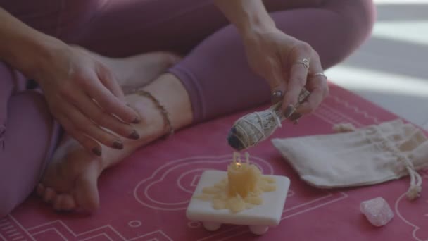 Niña yogui caucásica sentada en un yoga rosa mate, iluminando las hojas de salvia de una vela para la terapia del aroma. Usando leggins rosados. Todavía cerca de vídeo. — Vídeo de stock