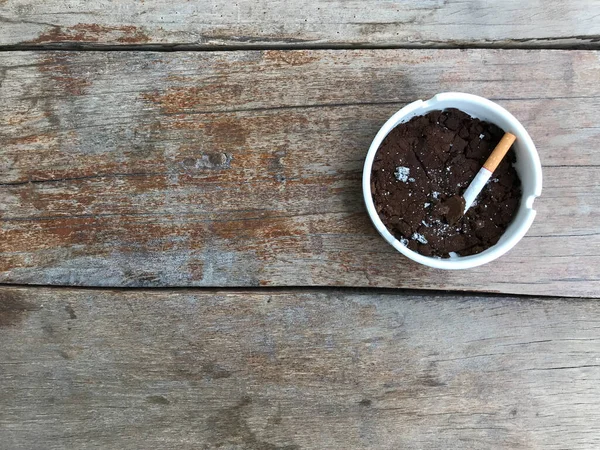 a smoked cigarette butt in a white ceramic ashtray which is full of used coffee grounds on a natural rough wood table with copy space on the left side of the frame