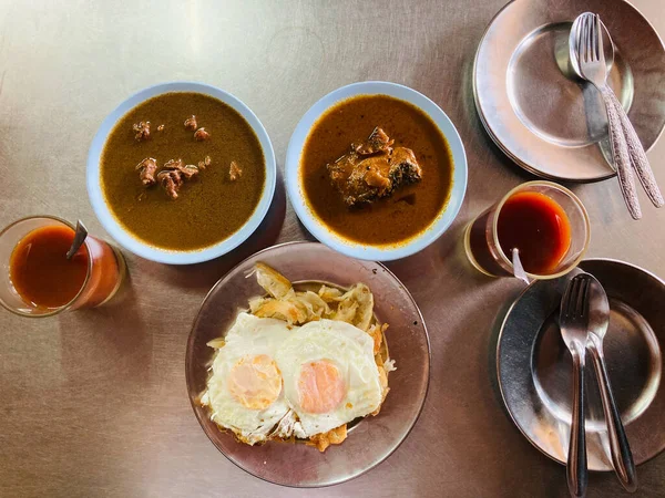 Phuket, Thailand breakfast style which has a bowl of fish and meat curry soup, a dish of Roti and two sunny side up or fried eggs, and two glasses of hot Ceylon tea, put on a stainless steel table