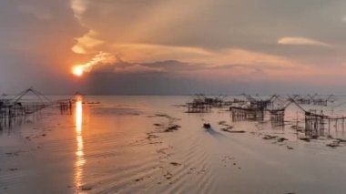 the boat is slowly sailing into the middle of golden sunrise light making wide amazing waves on the clear sea surface with the big square dip fish-trap net view