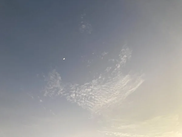 tiny crescent moon with an amazing shape of light white clouds on the pale grey-blue sky in natural daylight