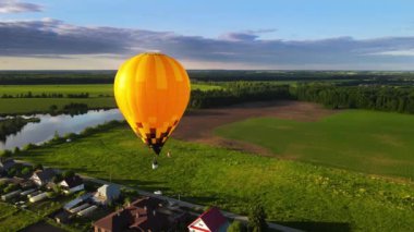 An orange air balloon above the field glows in dazzling rays of the setting sun. Aeronauts leisurely fly over the village on river bank. A picturesque and peaceful landscape at the evening golden hour
