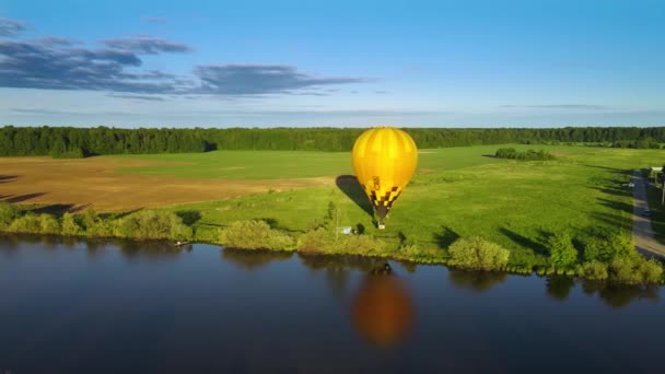 巨大な黄色の気球は 夕日の光線で川の岸の上にゆっくりと上昇します 気球のエンベロープは 水に反映されます ゴンドラには気球乗りが含まれています 商業航空業務 — ストック動画