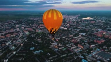 Sıcak hava balonu akşam manzarası üzerinde uçar. Parlak turuncu bir zarf karanlık bir şehre karşı parlıyor. Gondolda insanlar var. Pilot havayı propan meşalesiyle ısıtıyor. Dramatik bulutlu Ufuk.