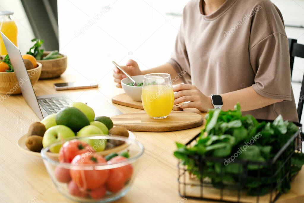 Woman eating fresh salad in kitchen and looking at laptop screen. Healthy lifestyle, healthy food, fresh juice. 