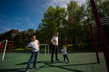 Beyaz bir adam ve iki çocuk dışarıda egzersiz yapıyor. Baba ve oğulları spor sahasında antrenman yapıyor.
