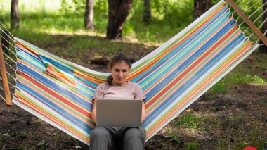 Ormandaki bir hamakta otururken dizüstü bilgisayarda çalışan beyaz bir kadın. Yürüyüşte kablosuz bilgisayar kullanan bir kız..