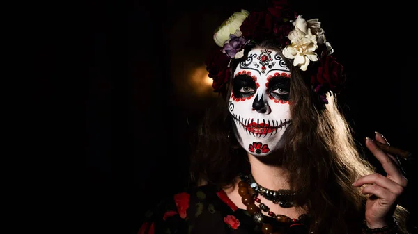 Mulher em Santa Muerte maquiagem em um fundo preto. Halloween menina fumar um charuto vestido como uma morte santa mexicana tradicional. — Fotografia de Stock