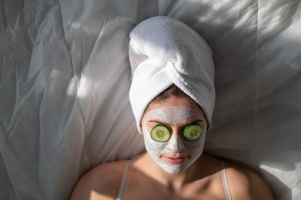 Una mujer con una toalla en el pelo y en una mascarilla de barro y pepinos frente a sus ojos yace sobre la sábana — Foto de Stock