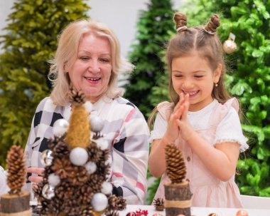 Yaşlı beyaz kadın iki torunuyla Noel için çam kozalağı süslemesi yapıyor.
