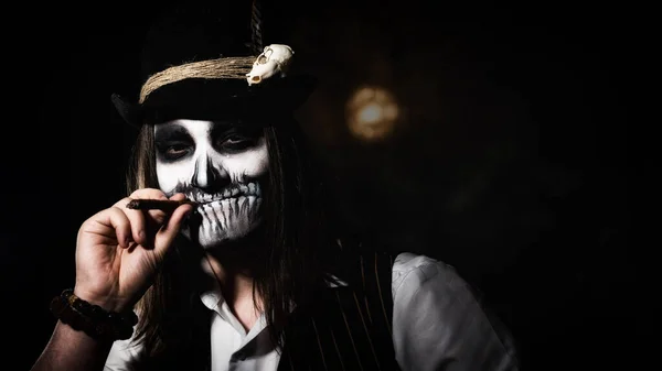 Um jovem de maquilhagem de crânio fuma um charuto no escuro. Halloween traje esqueleto — Fotografia de Stock