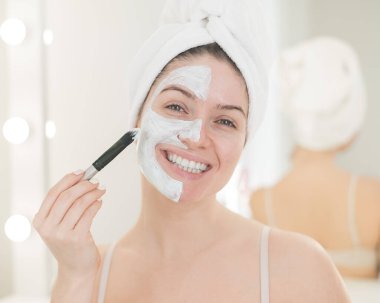 Saçında havlu olan güzel beyaz kadın kilden bir maske takıyor. Evdeki güzellikle ilgilenmek.