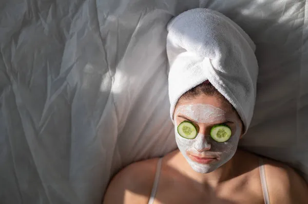 Una mujer con una toalla en el pelo y en una mascarilla de barro y pepinos frente a sus ojos yace sobre la sábana — Foto de Stock