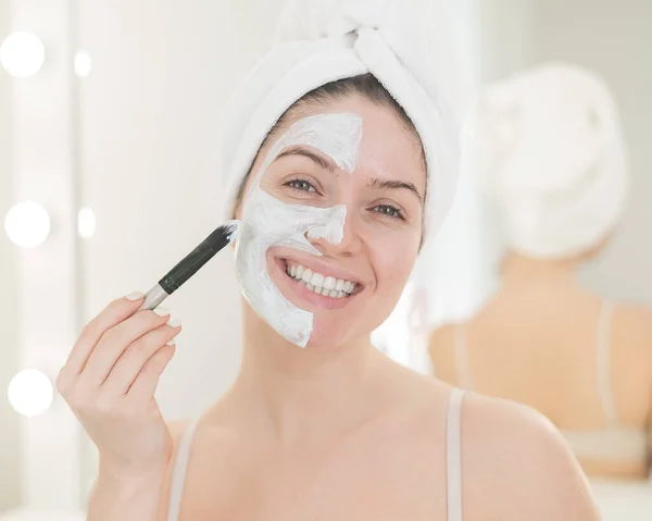 Beautiful caucasian woman with a towel on her hair applies a clay face mask. Taking care of beauty at home — Stock Photo, Image