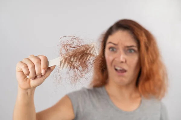 Mulher caucasiana com um sorriso de horror segura um pente com um pão de cabelo. Perda de cabelo e alopecia feminina. — Fotografia de Stock