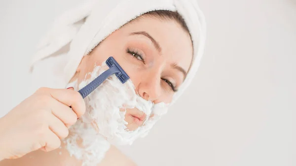 Веселая белая женщина с полотенцем на голове и пеной для бритья на лице держит бритву на белом фоне — стоковое фото