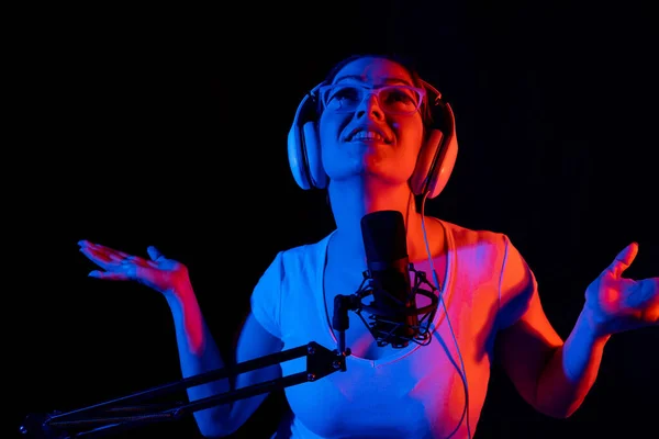 Mulher branca em óculos e fones de ouvido canta em um microfone em luz de néon em um fundo preto. Uma garota emocional está gravando uma música em um estúdio de gravação — Fotografia de Stock