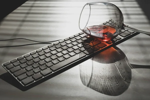 Una copa de vino tinto se encuentra en el teclado sobre una mesa blanca con un tono de las persianas — Foto de Stock