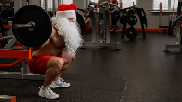 Santa claus in the gym. Muscular man with a naked torso doing exercises with dumbbells. — Stock Photo, Image