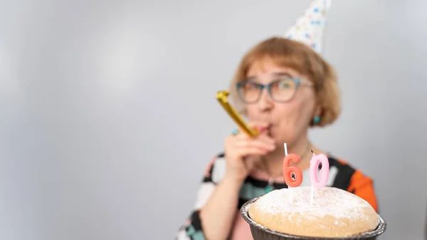 A portrait of a smiling elderly woman in a festive cap holding a cake with candles in the form of the number 60. Anniversary