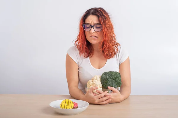 Kaukasisk kvinna som drömmer om snabbmat. rödhårig flicka väljer mellan broccoli och munkar på vit bakgrund. — Stockfoto