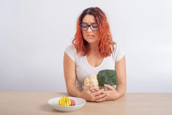 Femme caucasienne sur un régime rêvant de restauration rapide. rousse fille choisit entre brocoli et beignets sur fond blanc. — Photo