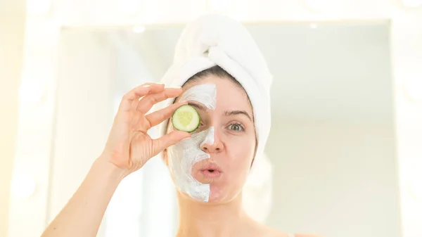 Radosna kobieta z ręcznikiem na włosach i w glinianej masce do twarzy zabawiająca się z ogórkami w dłoniach. Dbanie o piękno w domu — Zdjęcie stockowe