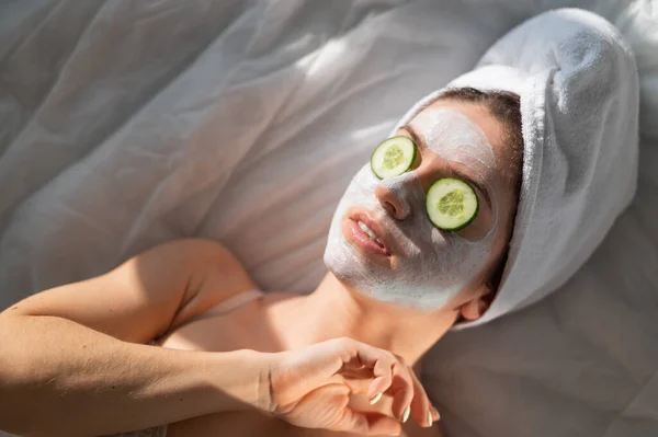 Una mujer con una toalla en el pelo y en una mascarilla de barro y pepinos frente a sus ojos yace sobre la sábana — Foto de Stock